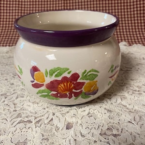 Peut inclure: Un bol rond en céramique avec une base de couleur crème et un bord violet foncé. L'extérieur présente un motif floral peint à la main avec des pétales et des feuilles rouges, jaunes et verts. L'intérieur est lisse et de couleur crème.