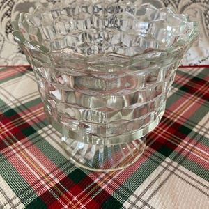 Puede incluir: Un cuenco de vidrio transparente con un patrón geométrico texturizado. El cuenco tiene un borde ancho y festoneado y se asienta sobre una base corta de pedestal. El cuenco se muestra sobre un mantel a cuadros con colores rojo, verde y blanco.