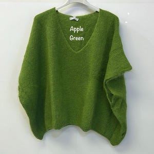 May include: An apple green knitted poncho with a V-neckline and a loose, relaxed fit. The garment features a soft, fuzzy texture and is displayed on a white hanger. The text Apple Green is printed in white on the front of the knitted fabric.