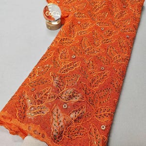 Könnte beinhalten: Orangener Spitzenstoff mit Blattmuster und kleinen Verzierungen. Der Stoff ist gefaltet und mit einem kleinen dekorativen Gegenstand präsentiert. Die Spitze hat ein detailliertes Design und eine strukturierte Optik, geeignet für Kleidung oder Bastelprojekte.