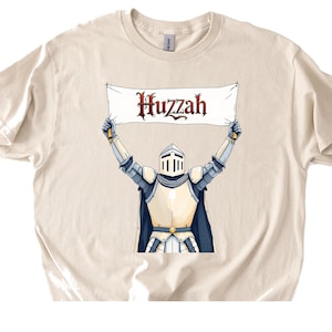 May include: Cream-colored t-shirt featuring a medieval knight illustration. The knight, clad in armor, holds a banner aloft with the word "Huzzah" in a decorative font. The design is centered on the shirt.