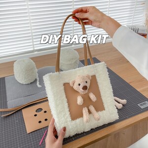 May include: A DIY bag kit featuring a fluffy white tote bag with a teddy bear applique. The bag has brown leather handles and a brown square patch with the bear. The words "DIY BAG KIT" are across the top.