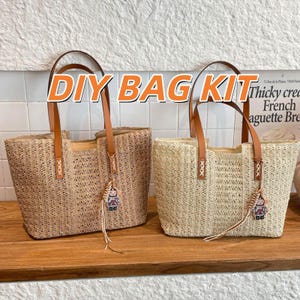 May include: Two woven tote bags with brown leather handles and decorative charms. The bags are made of a light brown material and have the text "DIY BAG KIT" in orange. The bags are sitting on a wooden surface.