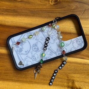 Könnte beinhalten: Schwarze Handyhülle mit weißem Totenkopf- und Knochenmuster. Die Hülle hat einen Perlenanhänger mit dem Text "OTMF" und einen weiteren Anhänger mit dem Text "BUNNY BAD". Der Anhänger enthält verschiedene bunte Perlen und silberfarbene Metallakzente.