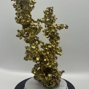 May include: A gold-colored, abstract sculpture with a porous, organic texture. The sculpture is mounted on a black and white circular base, creating a striking contrast. The intricate details and irregular shapes give it a unique, artistic appearance.