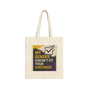 Puede incluir: Bolso tote de color crema con un diseño gráfico rectangular. El diseño gráfico muestra el texto "MY GENDER DOESN'T FIT YOUR CHECKBOX" en amarillo y morado, con un símbolo de verificación. El bolso tiene dos asas.