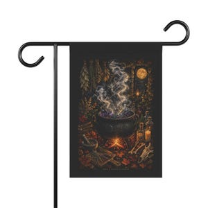 May include: A rectangular garden flag hanging on a black metal stand. The flag features a dark illustration of a black cauldron over a fire with rising smoke, surrounded by herbs, candles, glass bottles, a mortar and pestle, and a skull.