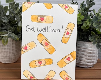 May include: A white greeting card with the words "Get Well Soon!" and illustrations of orange bandages with red heart centers. The card is displayed on a wooden surface with green plants in the background.