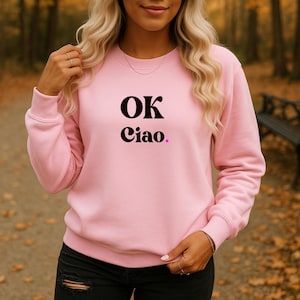 Könnte beinhalten: Ein hellrosa Sweatshirt mit dem Text "OK Ciao." in Schwarz und Lila. Das Sweatshirt hat einen Rundhalsausschnitt und besteht aus einem weichen Material. Das Model trägt eine schwarze Jeans.