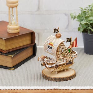 Könnte beinhalten: Ein Holzmodellschiff mit Totenkopf- und gekreuzten Knochen-Design auf dem Segel, auf einem Holzsockel. Das Schiff hat mehrere schwarze Flaggen und rot-weiß gestreifte Segel. Eine Sanduhr und Bücher befinden sich im Hintergrund.