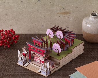 Peut inclure: Maquette en bois d'un sanctuaire japonais avec des portes torii rouges, des cerisiers en fleurs et des statues de renards. Le modèle est sur une base à plusieurs niveaux avec des éléments verts et beiges. Un vase décoratif et des baies rouges sont en arrière-plan.