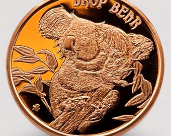 May include: A copper-colored coin featuring a detailed relief of a koala bear clutching eucalyptus leaves. The coin's rim is raised, and the words "DROP BEAR" are arched across the top. The coin has a shiny, reflective surface.