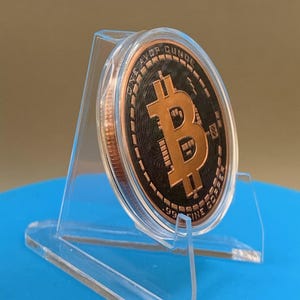 A copper-colored Bitcoin round encased in a clear plastic protective capsule, displayed on a transparent acrylic stand. It features a central Bitcoin symbol, with the text ONE AVOIRDUPOIS OUNCE and .999 FINE COPPER inscribed.