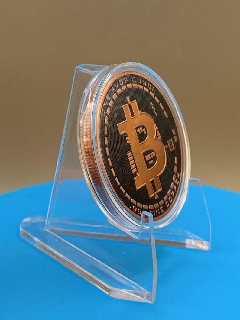 A copper-colored Bitcoin round encased in a clear plastic protective capsule, displayed on a transparent acrylic stand. It features a central Bitcoin symbol, with the text ONE AVOIRDUPOIS OUNCE and .999 FINE COPPER inscribed.