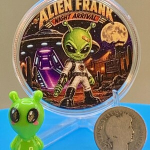 Night Arrival 1 oz copper round standing in its capsule beside a small green alien figurine and a silver coin for size comparison