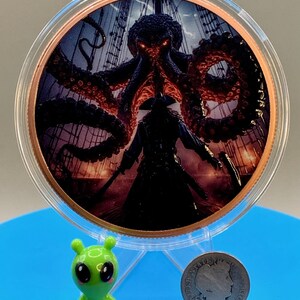 Puede incluir: Una moneda circular de tono cobrizo con una ilustraci&oacute;n colorida de un pulpo oscuro con ojos rojos brillantes sobre una figura pirata en la cubierta de un barco. Una peque&ntilde;a figura de alien&iacute;gena verde y una moneda de diez centavos Barber de 1906.