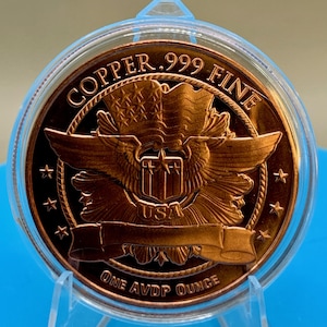 Close-up reverse view of a 1 oz .999 fine copper round showing an eagle crest with a shield and banner, "COPPER .999 FINE" and "ONE AVDP OUNCE" text, standing in a display holder on a blue background.