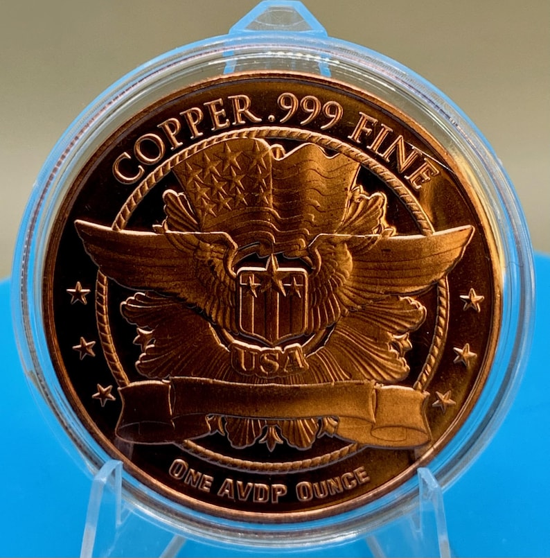 Close-up reverse view of a 1 oz .999 fine copper round showing an eagle crest with a shield and banner, "COPPER .999 FINE" and "ONE AVDP OUNCE" text, standing in a display holder on a blue background.