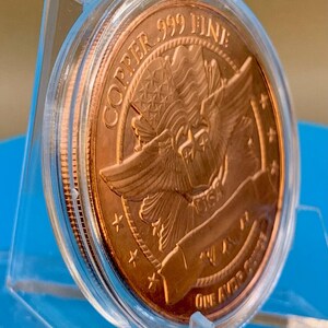 Angled edge-on view of a 1 oz .999 fine copper round in its display capsule, showing the round's thickness and reeded-style rim, standing on a blue background.