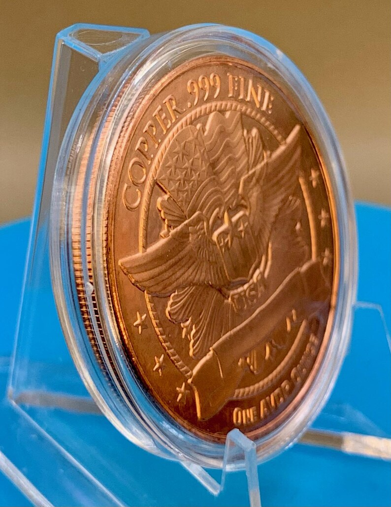 Angled edge-on view of a 1 oz .999 fine copper round in its display capsule, showing the round's thickness and reeded-style rim, standing on a blue background.