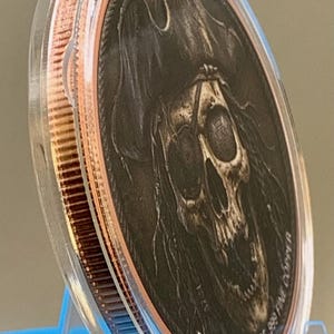 Puede incluir: Una moneda de cobre redonda encerrada en una c&aacute;psula protectora de pl&aacute;stico transparente. La moneda presenta un relieve detallado de una calavera con sombrero de pirata. El texto 5 AVDP OUNCES OF .999 FINE COPPER est&aacute; grabado en el borde.