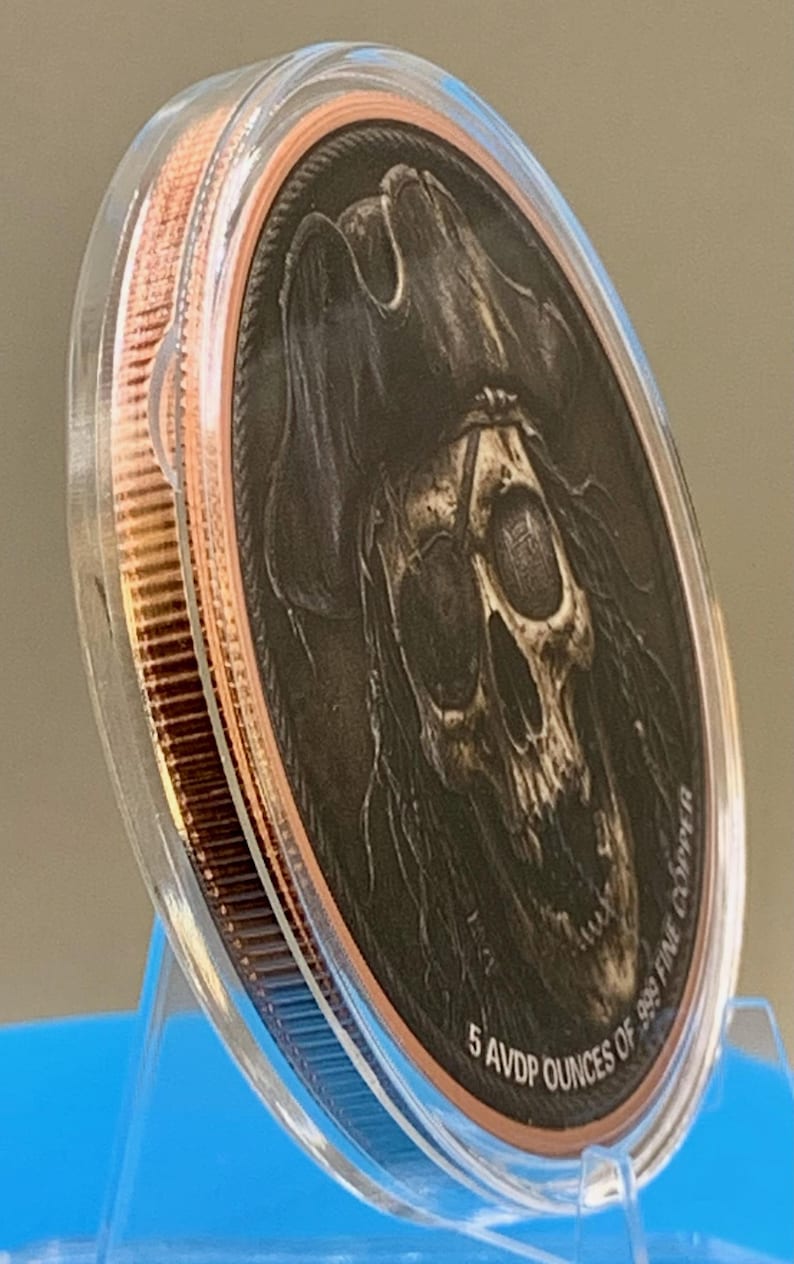 Puede incluir: Una moneda de cobre redonda encerrada en una c&aacute;psula protectora de pl&aacute;stico transparente. La moneda presenta un relieve detallado de una calavera con sombrero de pirata. El texto 5 AVDP OUNCES OF .999 FINE COPPER est&aacute; grabado en el borde.