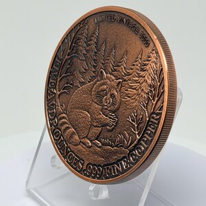 May include: A round copper coin featuring a relief of a raccoon sitting in a forest. The text reads LIMITED MINTAGE 5000 and FIVE AVDPOUNCES .999 FINE COPPER. The coin has a reeded edge and is displayed at an angle, showing the detailed metal texture.