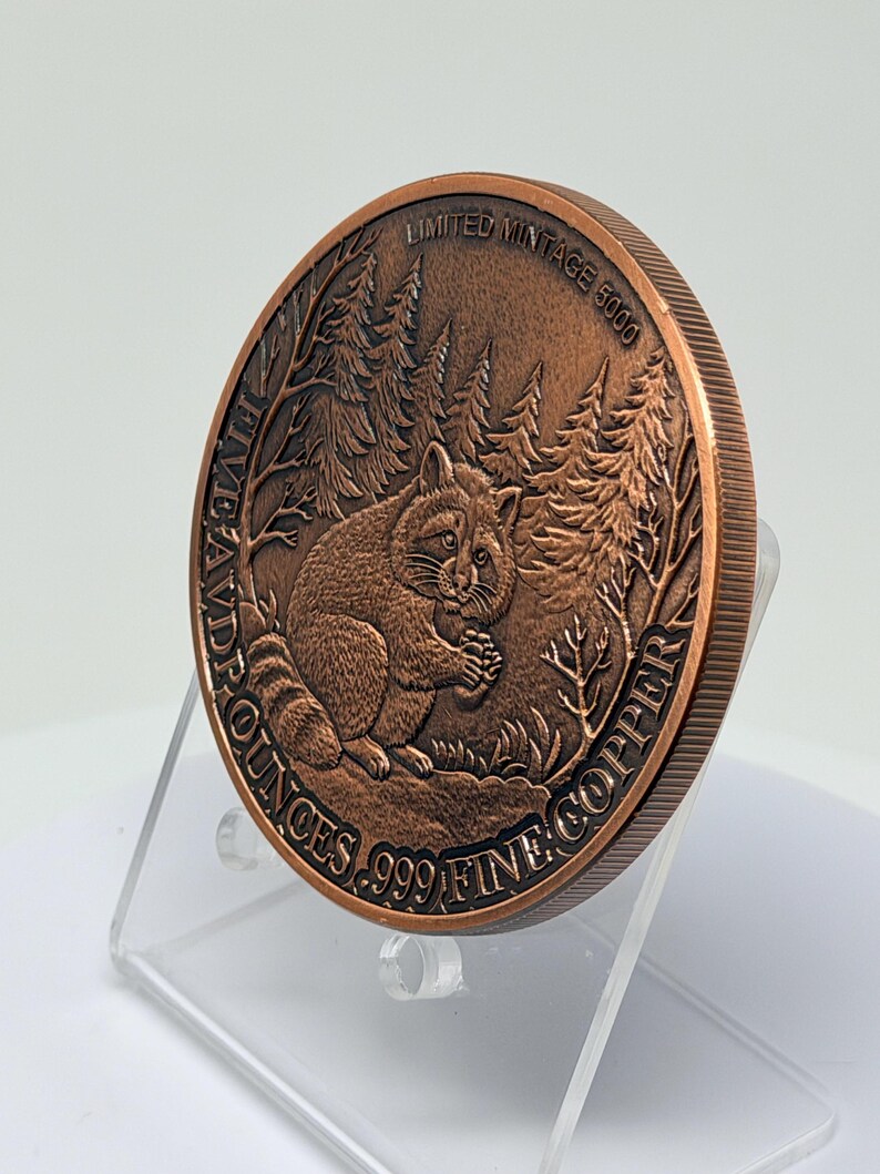 May include: A round copper coin featuring a relief of a raccoon sitting in a forest. The text reads LIMITED MINTAGE 5000 and FIVE AVDPOUNCES .999 FINE COPPER. The coin has a reeded edge and is displayed at an angle, showing the detailed metal texture.