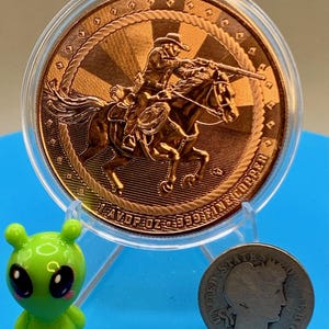 5 oz .999 fine copper Cowboy round shown with a small toy figure and a coin for size reference against a blue background.