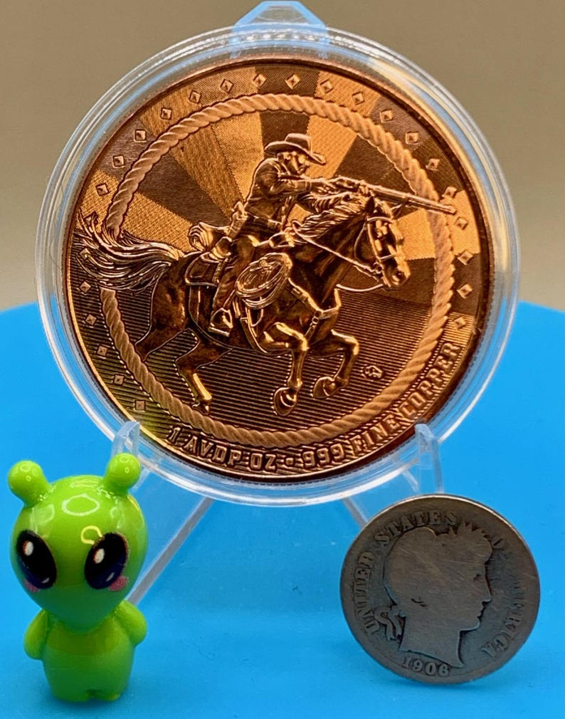 5 oz .999 fine copper Cowboy round shown with a small toy figure and a coin for size reference against a blue background.