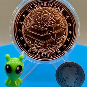 Reverse of the 1 oz copper round in a clear capsule, showing stacked copper bars with an atom and the Cu symbol and the words "Elemental" and "Stacker" around the rim, beside a small green alien figurine and an older silver-colored coin for scale.