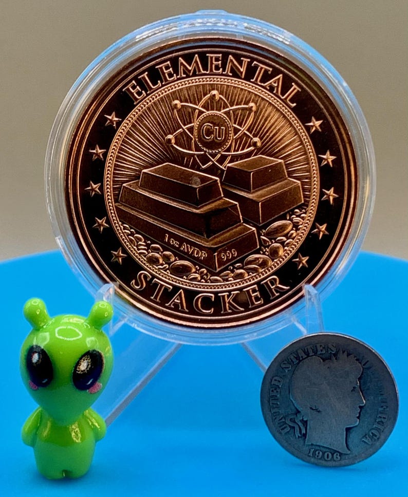 Reverse of the 1 oz copper round in a clear capsule, showing stacked copper bars with an atom and the Cu symbol and the words "Elemental" and "Stacker" around the rim, beside a small green alien figurine and an older silver-colored coin for scale.