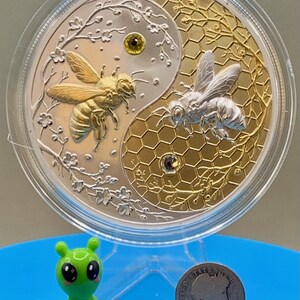 Harmony Bee copper round in its capsule shown with a small toy figurine and a US dime for scale reference, illustrating the round's large 5 oz size.