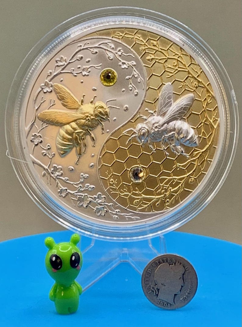 Harmony Bee copper round in its capsule shown with a small toy figurine and a US dime for scale reference, illustrating the round's large 5 oz size.