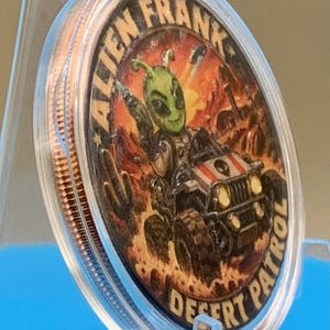 Angled side view of the Alien Frank Desert Patrol 1 oz copper round in its clear capsule on a blue stand, showing the colorized desert buggy artwork and the thick edge of the round.