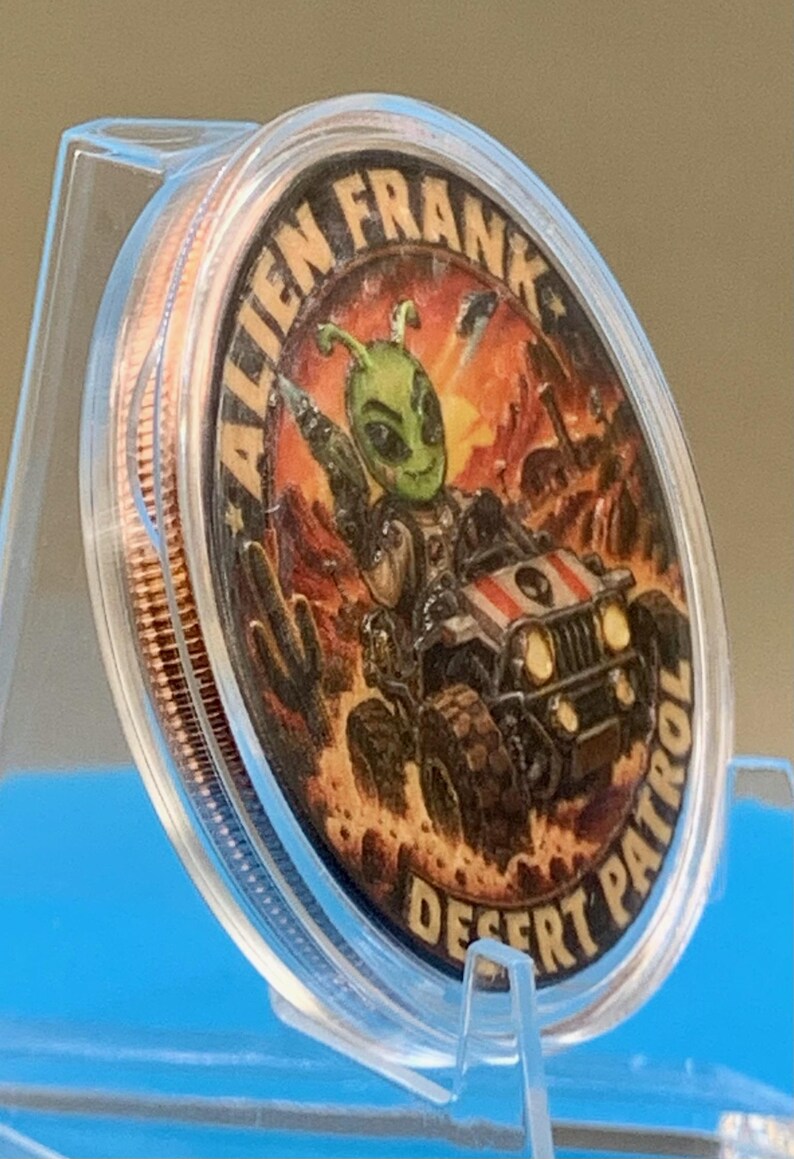 Angled side view of the Alien Frank Desert Patrol 1 oz copper round in its clear capsule on a blue stand, showing the colorized desert buggy artwork and the thick edge of the round.