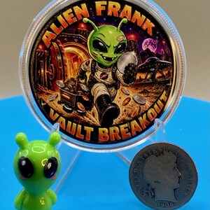 Alien Frank Vault Breakout 1 oz copper round in a clear capsule on a blue stand, shown next to a small green alien figurine and an older silver-colored coin to give a sense of scale.