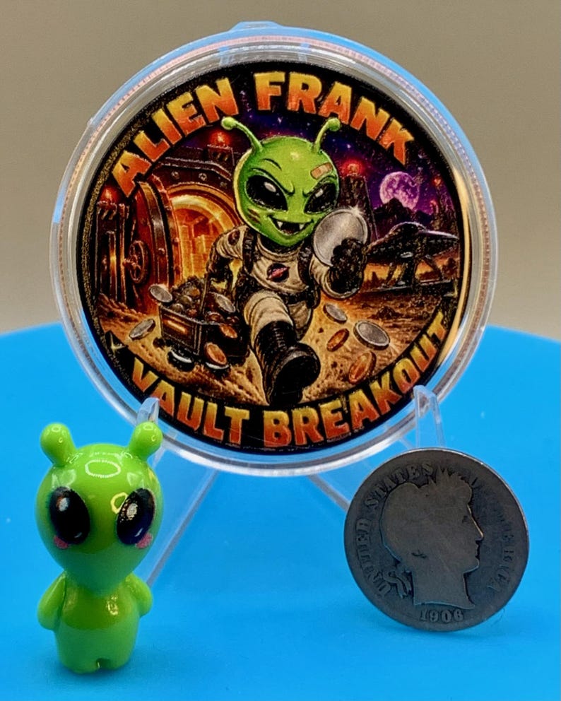Alien Frank Vault Breakout 1 oz copper round in a clear capsule on a blue stand, shown next to a small green alien figurine and an older silver-colored coin to give a sense of scale.