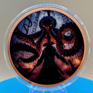 Puede incluir: Una moneda circular de tono cobrizo dentro de una c&aacute;psula protectora transparente, con un gr&aacute;fico a color de una criatura marina oscura de m&uacute;ltiples tent&aacute;culos con ojos rojos brillantes sobre una figura con abrigo en la cubierta.