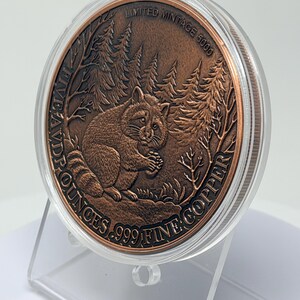 May include: A round copper coin encased in a clear plastic protective capsule. The coin features a relief of a raccoon in a forest setting. Text on the coin reads: LIMITED MINTAGE 5000, FIVE AVD P OUNCES, and .999 FINE COPPER.
