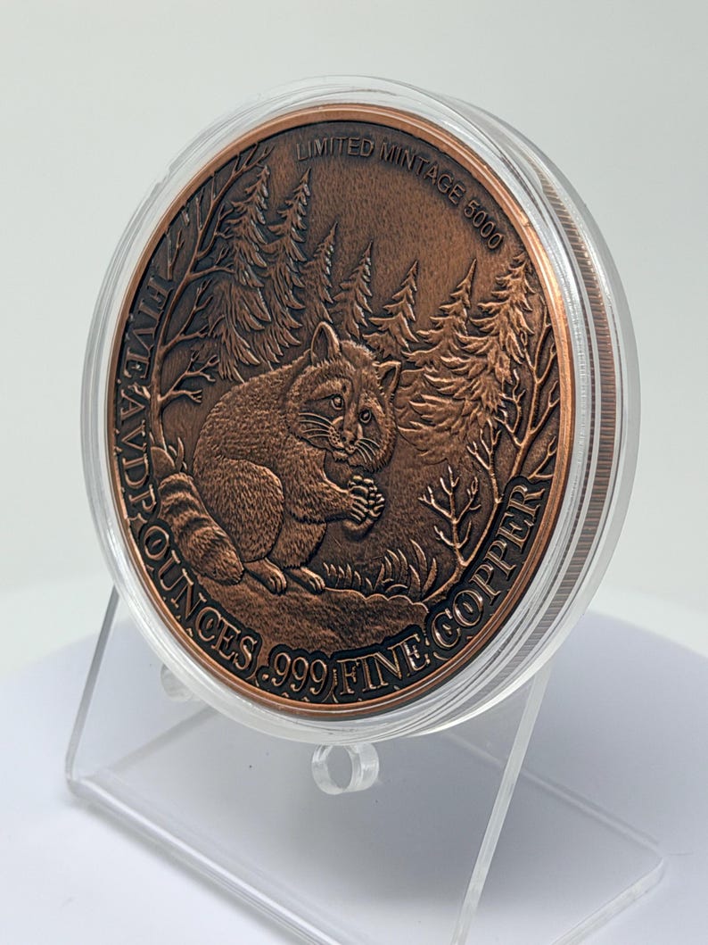 May include: A round copper coin encased in a clear plastic protective capsule. The coin features a relief of a raccoon in a forest setting. Text on the coin reads: LIMITED MINTAGE 5000, FIVE AVD P OUNCES, and .999 FINE COPPER.