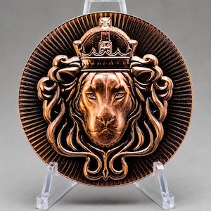 May include: A circular copper-toned medallion featuring a relief sculpture of a lion head wearing a crown. The lion is centered with a flowing mane, surrounded by a sunburst pattern radiating toward the edge of the coin-like object.