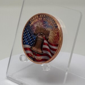 Angled side view of the colorized Liberty Bell and American flag 1 oz copper round on a clear acrylic stand, showing the coin's thickness and raised relief edge