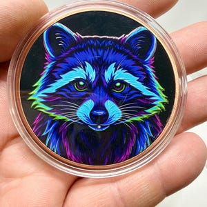 May include: A circular coin featuring a vibrant, neon-colored illustration of a raccoon face against a black background. The raccoon is rendered in shades of electric blue, purple, and bright green, encased in a clear protective plastic capsule.