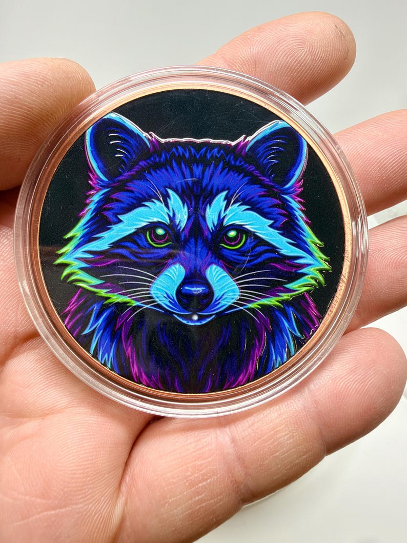 May include: A circular coin featuring a vibrant, neon-colored illustration of a raccoon face against a black background. The raccoon is rendered in shades of electric blue, purple, and bright green, encased in a clear protective plastic capsule.