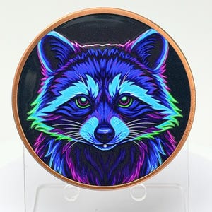 May include: A circular metal coin featuring a vibrant, stylized illustration of a raccoon face. The raccoon is rendered in neon shades of blue, purple, and green against a dark background, with a copper-colored metallic rim encircling the design.