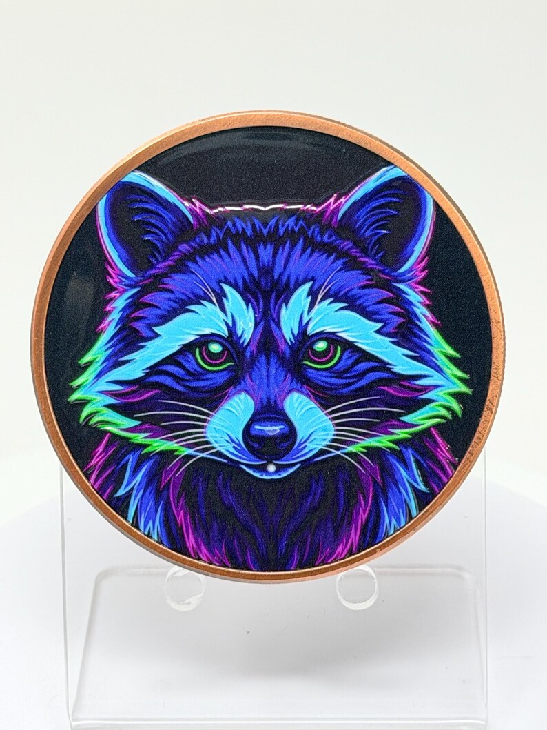 May include: A circular metal coin featuring a vibrant, stylized illustration of a raccoon face. The raccoon is rendered in neon shades of blue, purple, and green against a dark background, with a copper-colored metallic rim encircling the design.