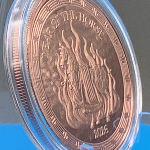 Angled view of the encapsulated 2026 Year of the Horse copper round tilted on a blue stand, showing the armored horse and flames obverse with Greek key border, the 2026 date and the reeded edge.