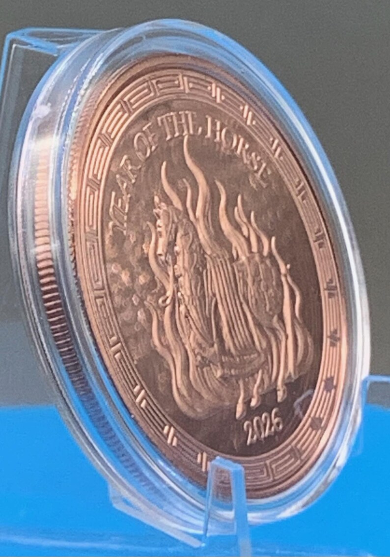 Angled view of the encapsulated 2026 Year of the Horse copper round tilted on a blue stand, showing the armored horse and flames obverse with Greek key border, the 2026 date and the reeded edge.