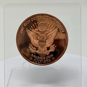 Reverse of the 1 oz .999 copper round showing the Great Seal eagle, E Pluribus Unum and July 4th, standing on a clear acrylic stand against a plain light background